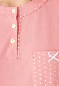 Camicia di cotone rosa con scollatura tondeggiante, quattro bottoni pesca e una tasca con pois bianchi e un nastro bianco decorativo.
