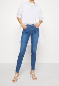 Žena stojí s rukama v kapsách, má na sobě modré úzké džíny, bílé sandály na klínu s pásky, bílé tričko a rozepnutou bílou košili.