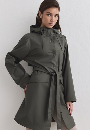 Donna che indossa un impermeabile verde scuro con cappuccio, cintura in vita e grandi tasche frontali, in piedi contro un muro bianco testurizzato.
