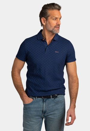 Man met baard die een marineblauw textuur poloshirt en lichtblauwe spijkerbroek draagt, staand met één hand in zijn zak, kijkend naar zijn rechterkant.