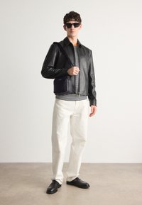Giacca di pelle nera, camicia a righe, pantaloni bianchi, scarpe nere. Tiene una borsa scura con un motivo sottile. Gli accessori includono occhiali da sole neri.