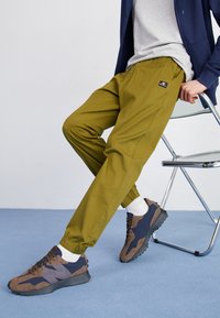 Olivgröna joggerbyxor med elastiska muddar, prydda med en logotyp; kombinerade med bruna och navy sneakers och vita strumpor.