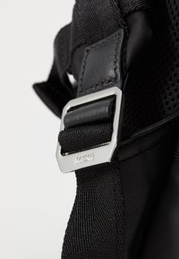 Detaliile unei genți din material textil negru, prezentând o cataramă rectangulară argintie cu logo-ul "MCM", un curea texturată și un panou din plasă în contrast.