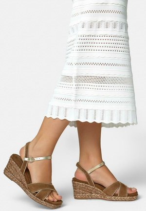 Piernas con sandalias de cuña marrones con correas doradas en el tobillo, combinadas con un vestido blanco hasta la rodilla con patrones horizontales de encaje.