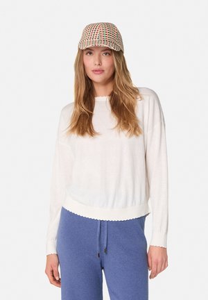 Witte lange mouw top met een gekartelde zoom, gecombineerd met blauwe joggingsbroek. De top heeft een lichte stof en een losse pasvorm.