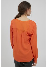 Oranges langärmliges Shirt mit rundem Ausschnitt, das ein gerafftes Rückendesign und eine glatte Textur aufweist. Kombiniert mit dunklen Hosen.