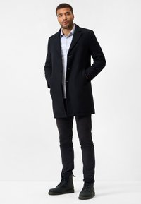 Black wool coat with a notched collar, single-breasted front, and button closure. Paired with dark pants and black lace-up boots.