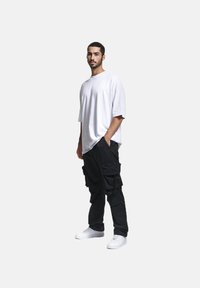 Hvit oversized T-skjorte kombinert med svarte cargo-bukser med flere lommer. Modellen står mot en ensfarget bakgrunn iført hvite sneakers.