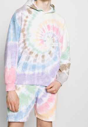 Persoon die een pastel tie-dye hoodie draagt en bijpassende shorts met spiraal patroon in roze, paars, groen, blauw en beige op witte achtergrond.