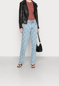 Schwarze Lederbikerjacke, bordeauxrote taillierte Bluse, hellblaue karierte Jeans und schwarze Sandalen. Das Model hält eine schwarze Handtasche.