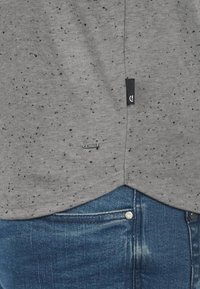 Grå t-shirt med et spættet mønster, lavet af blødt stof. Nederkanten er let buet med et sort stofmærke i siden. Båret med blå jeans.