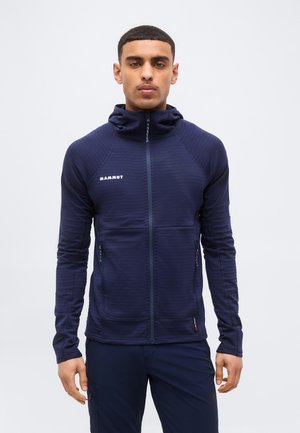 Mężczyzna w granatowej kurtce Mammut z zamkiem błyskawicznym, kapturem i kieszeniami na suwak, stojący na jasnym, jednolitym tle.