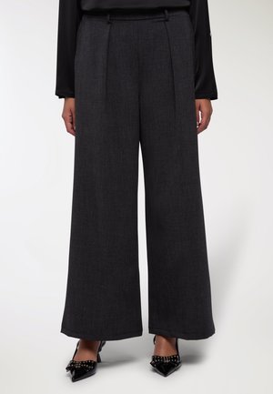 Pantaloni a gamba larga in tessuto grigio scuro, con vita alta, pieghe sartoriali e una texture liscia. Abbinati a tacchi neri a punta.