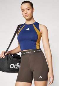 Débardeur sans manches bleu et doré avec un col rond et un logo ; short ajusté marron avec ceinture noire ; sac de sport noir avec logo.