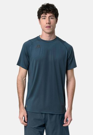 Giovane uomo con capelli ricci che indossa una maglietta sportiva a maniche corte color verde petrolio scuro e pantaloncini abbinati su uno sfondo bianco uniforme.