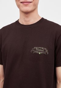 Νέος που φοράει σκούρο καφέ T-shirt Carhartt με μικρό κρεμ χρωματιστό διακοσμημένο λογότυπο στο αριστερό στήθος.