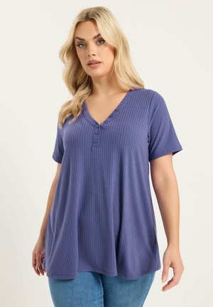 Frau trägt ein locker sitzendes, kurzärmeliges, lila, geripptes Oberteil mit V-Ausschnitt und drei Knöpfen, kombiniert mit blauen Jeans vor hellem Hintergrund.