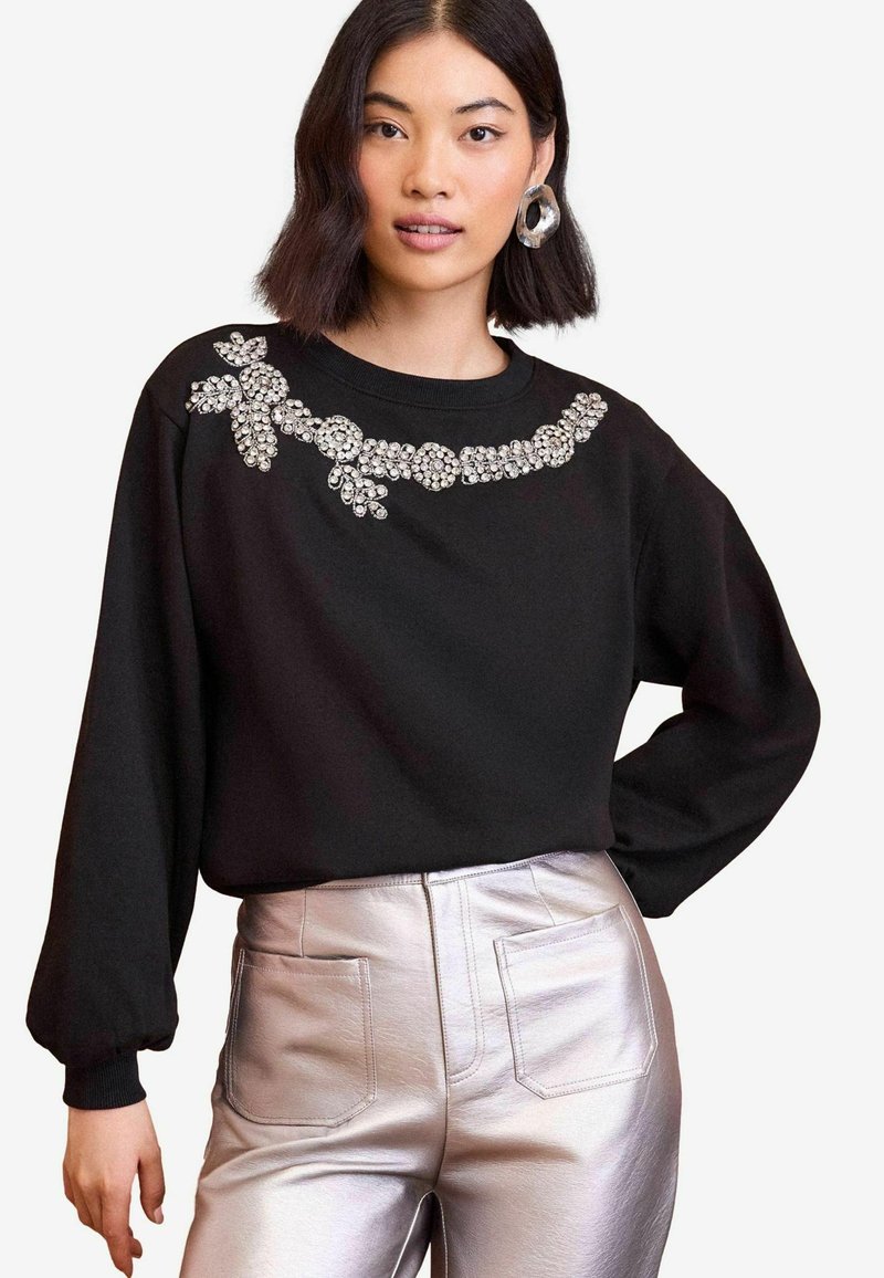 Crna dukserica s okruglim izrezom, ukrašena prozirnim rhinestones u cvjetnom dizajnu. Uparena s metalik srebrnim hlačama visokog struka.