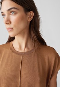 Braunes Kurzarmoberteil mit Rundhalsausschnitt und dezentem diagonalen Faltendetail. Weicher, elastischer Stoff mit glatter Textur.