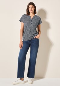 Marinblå mönstrad kortärmad blus med v-ringning och tre knappar, kombinerad med vida mörkblå jeans och vita sneakers.