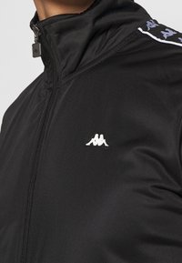 Chaqueta negra con cremallera hecha de una tela suave, que presenta un cuello alto y un logotipo blanco en el pecho, con detalles estampados en los hombros.
