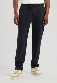 Donkerblauwe broek in een relaxte pasvorm, met een trekkoord in de taille, riemlussen, zakken en een gladde stof. Gepaard met lichtgekleurde sneakers.