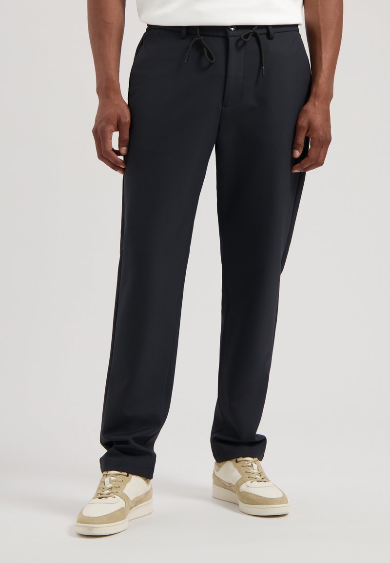 Donkerblauwe broek in een relaxte pasvorm, met een trekkoord in de taille, riemlussen, zakken en een gladde stof. Gepaard met lichtgekleurde sneakers.