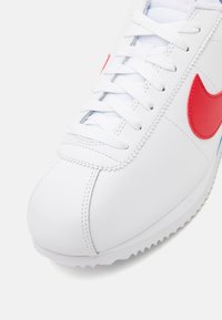 White leather sneaker with red Nike swoosh, textured upper, padded tongue, and flat laces. Rubber sole with zigzag pattern for traction.