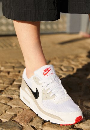 Personne portant des baskets Nike Air blanches avec un swoosh noir et des accents rouges, debout sur un pavé à côté d'une ombre.