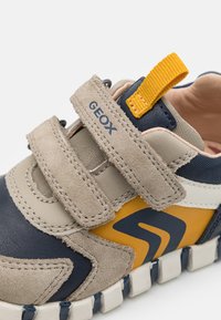 Velcro tenisky v námořnické modré, béžové a žluté barvě. Mají semišové a hladké kožené materiály, výrazné logo a teksturovanou gumovou podrážku.