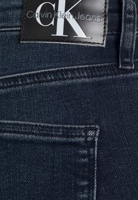 Tela de mezclilla azul oscuro con una textura sutil, que presenta una etiqueta de cuero negro que dice "Calvin Klein Jeans" con un logo plateado.