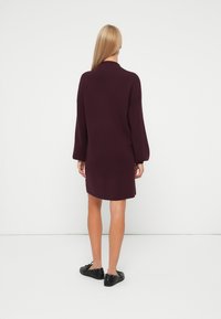 Vestito in maglia di un profondo bordeaux, con orlo leggermente arrotondato, maniche lunghe e vestibilità morbida. Indossato con scarpe nere con lacci.
