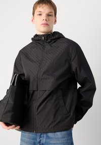Zwarte windjack met een geometrisch patroon, ritssluiting aan de voorkant en capuchon. Lichtgewicht stof met een gladde textuur en twee zijzakken.