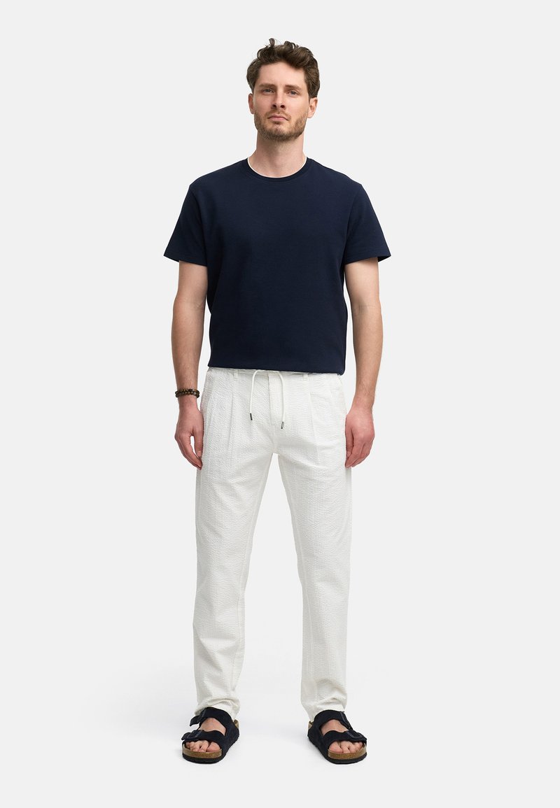 Marine bomulds-tshirt, hvide teksturerede bukser og sorte sandaler med stropdesign. Model står mod en ensfarvet hvid baggrund.