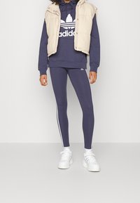 Námořnický sportovní sweatshirt s bílým logem, který je kombinován s odpovídajícími legínami s bočními pruhy a béžovým péřovým vestou. Bílé tenisky dokončují vzhled.