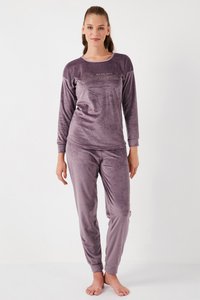 Joven de pie descalza, vistiendo un conjunto de terciopelo morado con mangas largas y pantalones, con el texto "Sigue tus sueños" en el pecho.