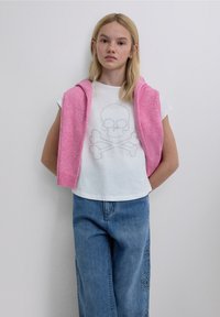 Weißes Kurzarmhemd mit Totenkopf- und Knochen-Design, kombiniert mit hellblauen Jeans und einem pinkfarbenen Pullover, der über die Schultern gelegt ist.