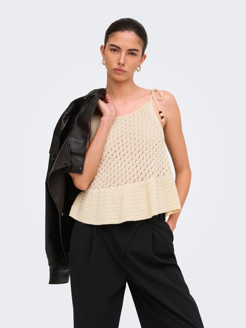 Femme portant un haut sans manches en maille beige avec un ourlet à volants, un pantalon noir, des boucles d'oreilles créoles dorées, tenant une veste en cuir noire sur son épaule.