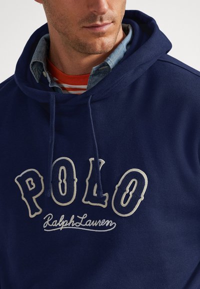 Vyras, dėvintis sluoksniuotą tamsiai mėlyną gobtuvą su ant priekinės dalies išsiuvinėtu užrašu „POLO Ralph Lauren“ ir po juo mėlyną mygtukais užsegamą marškinių palaidinę.