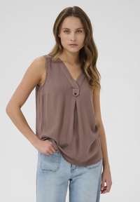 Bruine mouwloze top met een V-hals en een klein knoopdetail aan de voorkant. De stof heeft een gestructureerde afwerking, gecombineerd met lichtblauwe jeans.