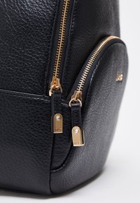 Sort læderrygsæk med tekstureret overflade, gyldne lynlåse og en rundet sidelomme med brandede hardware-accenter.