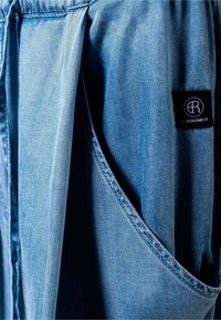 Φωτεινό γαλάζιο denim ύφασμα με ορατή ραφή και μαύρη ορθογώνια ετικέτα μάρκας στην δεξιά πλευρά που γράφει "ELIAS RUMELIS".