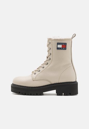 Stivale da combattimento in pelle beige con finitura testurizzata, allacciatura frontale, collo foderato in pelliccia e suola carrarmato nera; presenta una toppa con logo sul lato.