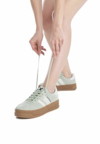 Zapatillas de ante verde con rayas blancas, punta redondeada y suela gruesa de goma marrón. Se están atando los cordones con las manos en primer plano.