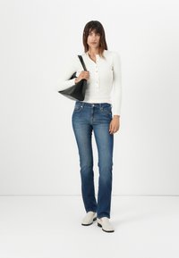 White long-sleeve henley top with buttons, blue denim jeans, and cream shoes. Black shoulder bag. Standing against a white backdrop.
