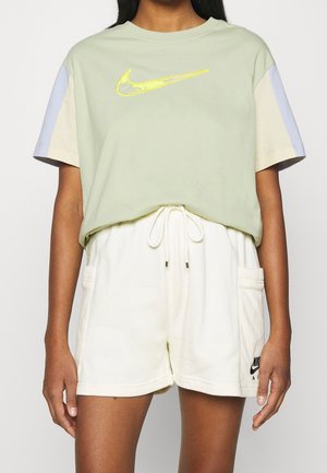 Osoba ubrana w pastelowo zieloną koszulkę Nike z żółwym logo Swoosh oraz białe szorty ze sznurkiem i kieszeniami, z czarnym logo Nike Air na lewej nodze.