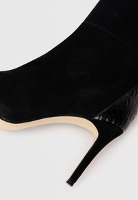 Bottine en daim noir avec un talon aiguille, dotée d'un accent en cuir verni noir texturé sur le côté. Surface lisse et design épuré.