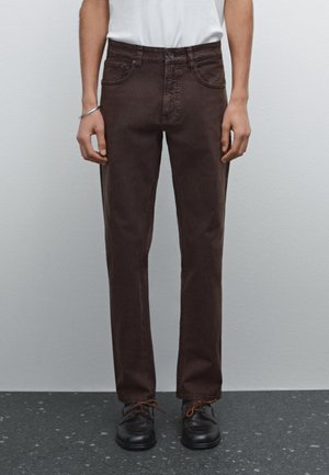 Persona che indossa jeans marrone scuro a gamba dritta, scarpe nere con lacci e una camicia bianca, in piedi contro un muro grigio chiaro uniforme.