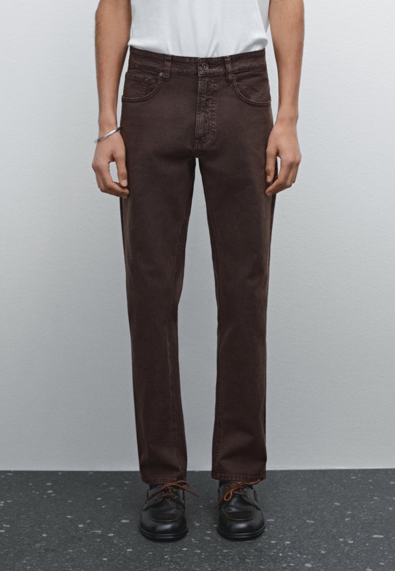 Personne portant un jean droit marron foncé, des chaussures noires à lacets et une chemise blanche, debout contre un mur gris clair uni.