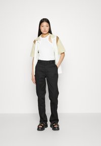 White tank top with cutouts, layered with a light green and brown short-sleeve shirt, paired with high-waisted black trousers and chunky sandals.